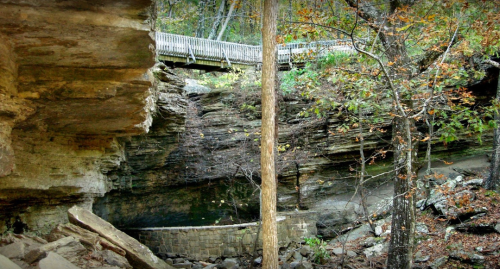 A rocky landscape with a wooden bridge above, surrounded by trees and autumn foliage.