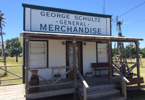 A small, rustic general store with a sign reading "George Schultz General Merchandise" under a clear blue sky.