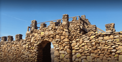 Stone ruins with a wooden railing against a clear blue sky, featuring an arched entrance and weathered stones.