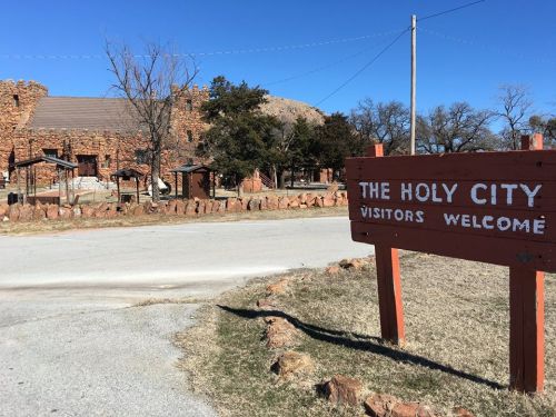 Sign reading "The Holy City, Visitors Welcome" in front of a stone building and trees along a rural road.