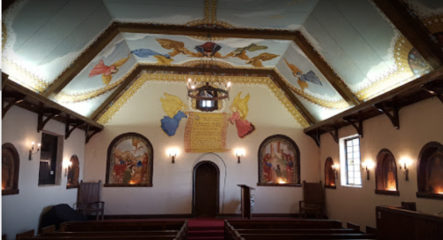 Interior of a chapel featuring ornate ceiling murals, angelic figures, and wooden pews, creating a serene atmosphere.
