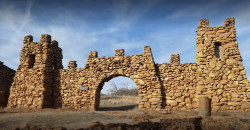 Stone archway with towers, made of large rocks, set against a blue sky and grassy landscape.