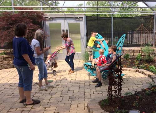 A group of adults and children in a garden, with colorful butterfly chairs and plants around them.