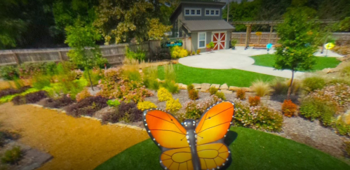 A colorful garden featuring a large butterfly sculpture, vibrant plants, and a shed in the background.