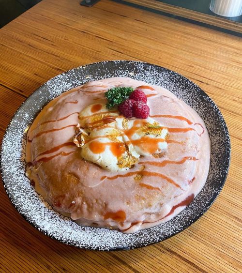 A large, fluffy pancake topped with syrup, raspberries, and a dollop of cream, served on a black plate.