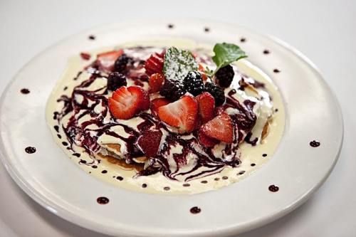 A plate of pancakes topped with strawberries, blackberries, cream, and a berry sauce drizzle.