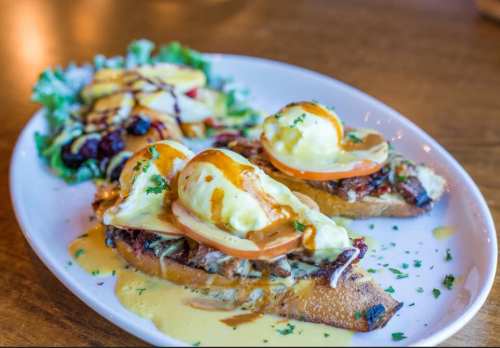 A plate of eggs Benedict on toasted bread, topped with hollandaise sauce, served with a side salad and fruit.