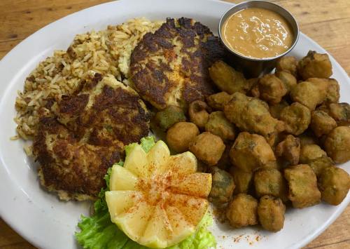 A plate of rice, two crab cakes, fried okra, and a small cup of sauce, garnished with lettuce and pineapple.