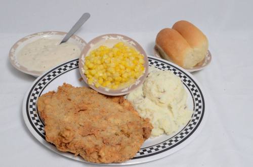 A plate of fried chicken, mashed potatoes, corn, and two dinner rolls, with a side of gravy in a bowl.
