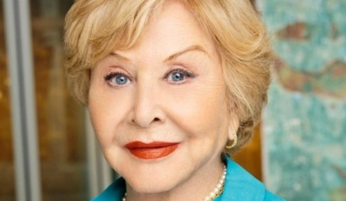 A smiling older woman with blonde hair and blue eyes, wearing a turquoise blouse and pearl earrings.