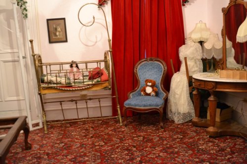 A vintage room with a brass bed, red curtains, a blue chair, a teddy bear, and elegant dresses displayed.