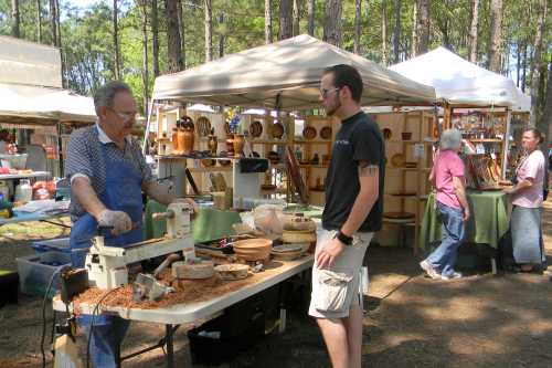 A craftsman demonstrates woodturning at a market, while a visitor observes. Stalls with wooden items are in the background.