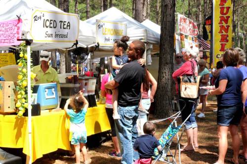 A busy outdoor market scene with people at food stalls selling iced tea and lemonade among tall trees.