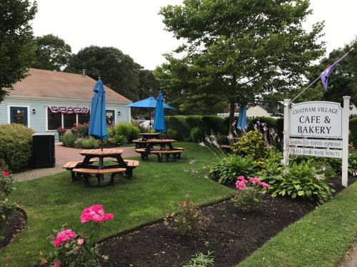 A quaint café and bakery with outdoor seating, surrounded by flowers and greenery, under blue umbrellas.