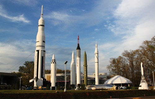 A collection of historic rockets and spacecraft displayed outdoors against a blue sky and trees.