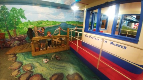 Children play on a wooden bridge in a colorful room with a mural of a river and a boat named "The Captain Jim Parker."