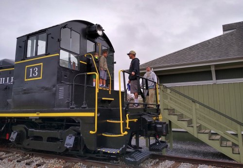 A group of people interacts near a vintage black train engine at a station, with stairs leading up to it.