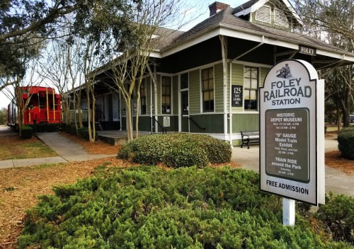 Foley Railroad Station with a sign for the museum and a red model train in the background, surrounded by greenery.