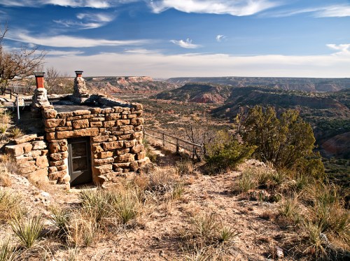 A stone structure overlooks a vast canyon landscape under a blue sky with wispy clouds.