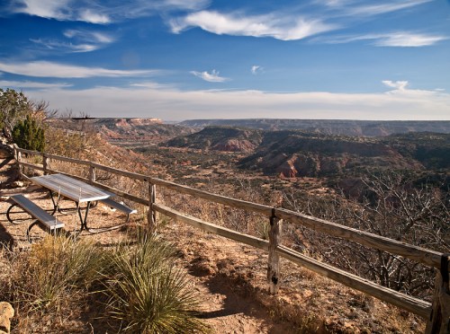 A scenic view of a canyon with a picnic table and wooden fence under a blue sky with wispy clouds.