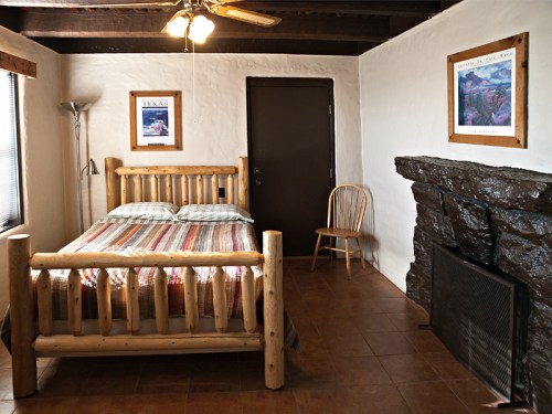 Cozy bedroom with a wooden bed, chair, and stone fireplace, featuring rustic decor and warm lighting.