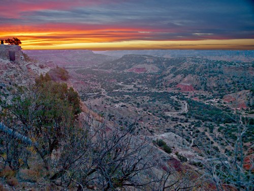 A vibrant sunset over a canyon landscape, with colorful skies and rugged terrain dotted with shrubs and trees.