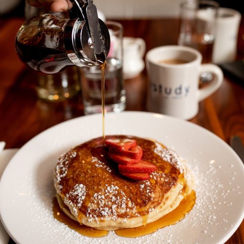 A stack of pancakes topped with strawberries, drizzled with syrup, served on a white plate with a coffee cup in the background.
