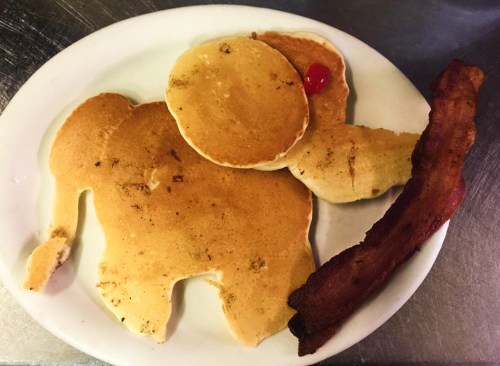 A plate with pancakes shaped like an elephant, topped with a cherry, alongside a strip of bacon.