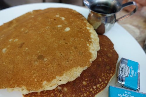 A plate with two golden-brown pancakes, a small metal syrup pitcher, and butter packets on the side.