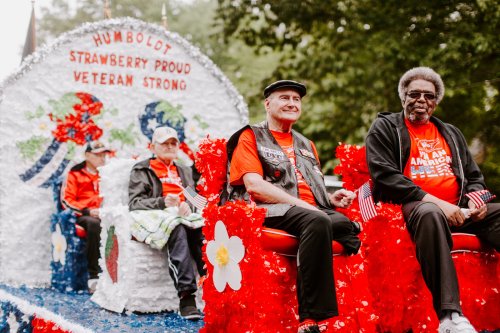 A festive parade float with veterans in red shirts, decorated with flowers and flags, celebrating community pride.