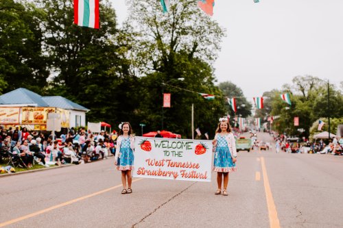 Two girls in blue dresses hold a banner reading "Welcome to the West Tennessee Strawberry Festival" at a parade.