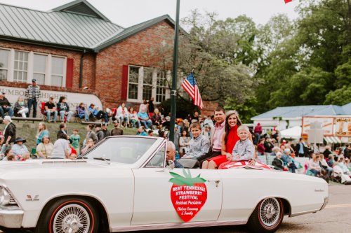 A convertible car in a parade with smiling adults and children, surrounded by a festive crowd and buildings.
