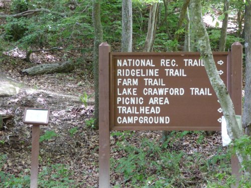 A brown trail sign in a wooded area, listing various trails and amenities like picnic area and campground.