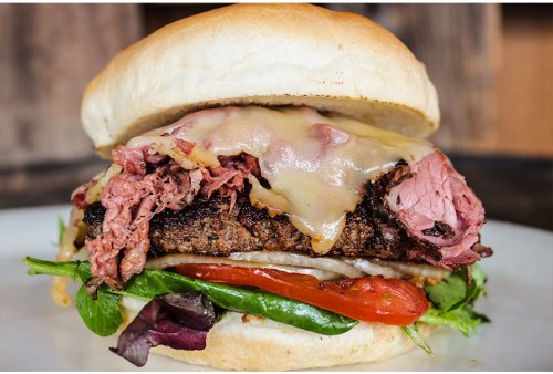 A close-up of a juicy burger with melted cheese, layers of meat, lettuce, tomato, and a soft bun on a plate.
