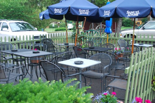 Outdoor seating area with black metal tables and chairs, shaded by blue umbrellas, surrounded by a white picket fence.