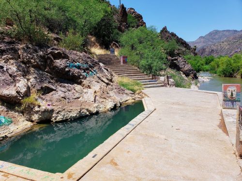 A rocky landscape with a clear water pool, steps leading up, and a river flowing nearby under a bright blue sky.