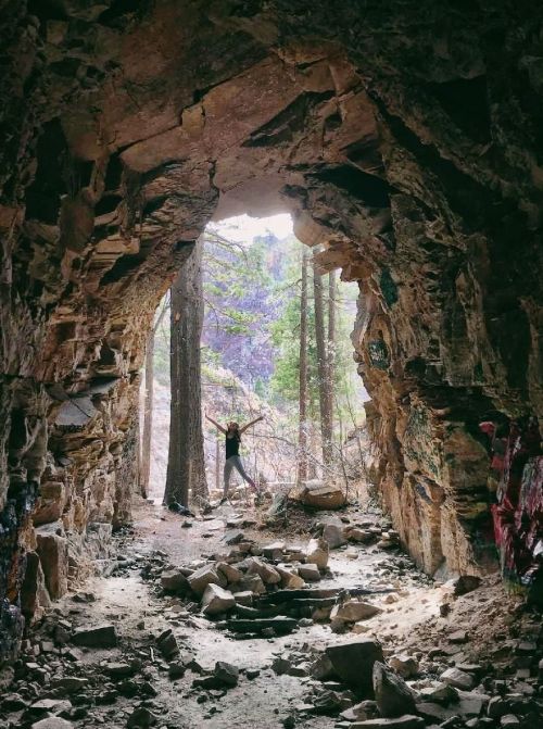 A person stands in a rocky cave entrance, surrounded by trees and natural light filtering through the opening.