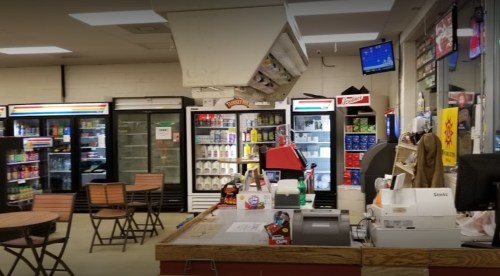 Interior of a convenience store featuring a checkout counter, shelves of drinks, and seating area with tables.