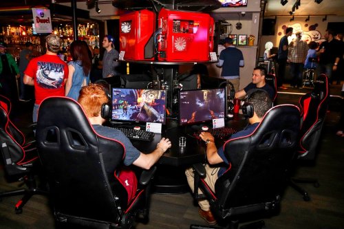 Two gamers sit at a circular gaming station with multiple screens, surrounded by a lively bar atmosphere.