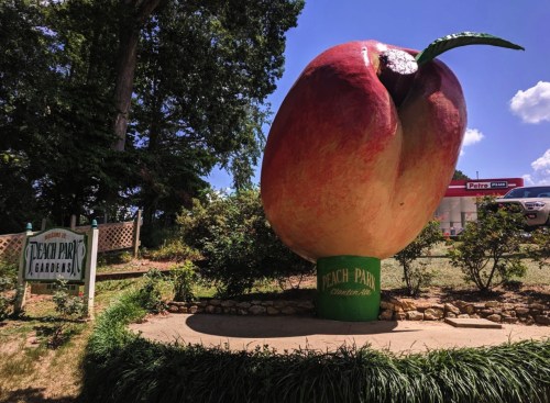 A large peach statue stands at the entrance of Peach Park Gardens, surrounded by greenery and a blue sky.