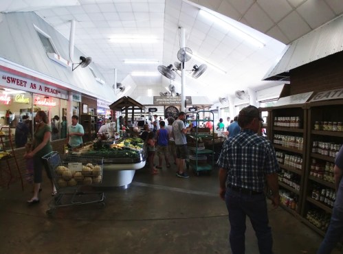 A bustling market interior with shoppers browsing fresh produce and goods, surrounded by shelves and displays.