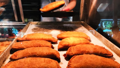 A tray of golden-brown empanadas with a hand tossing another empanada onto the tray.