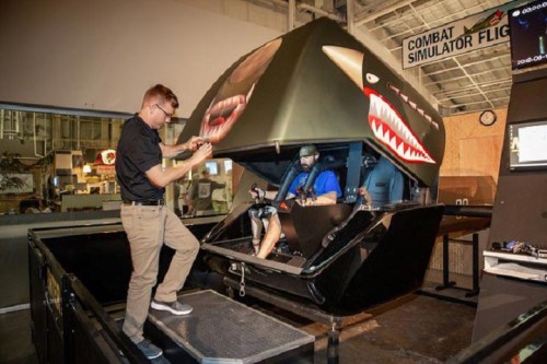 A person sits in a combat flight simulator while another adjusts controls outside the cockpit.