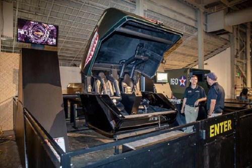 A military-themed simulator with two seats, surrounded by a display area and two men discussing it.