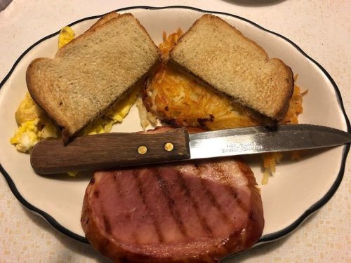 A plate with grilled ham, scrambled eggs, hash browns, and two slices of toasted bread, accompanied by a knife.