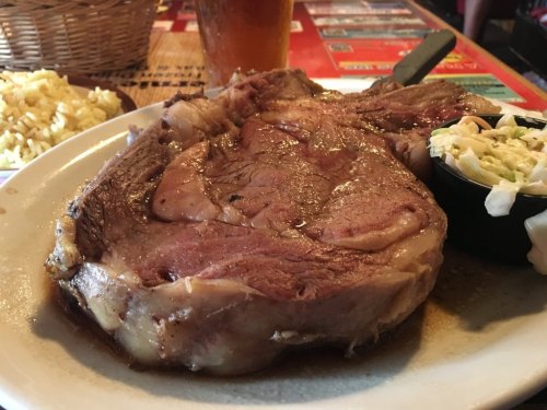 A large, juicy prime rib steak served with coleslaw and a side of macaroni, with a drink in the background.