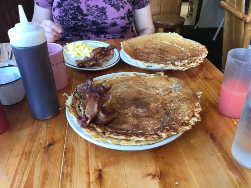 A table with two plates of large pancakes, bacon, and scrambled eggs, with drinks and condiments nearby.