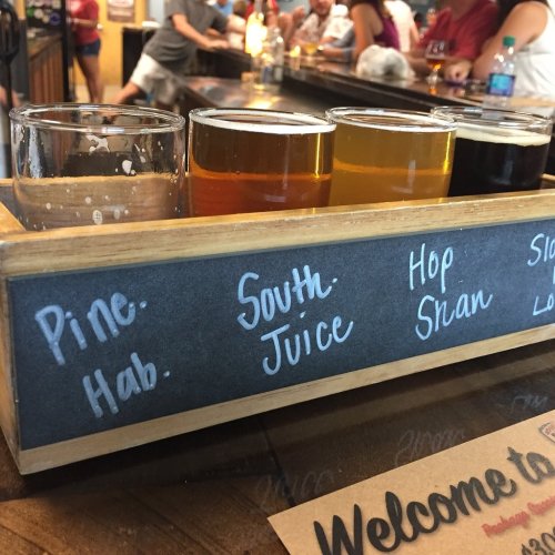 A wooden flight tray with four beer samples labeled Pine, Hab, South Juice, and Hop Shan, set on a bar.