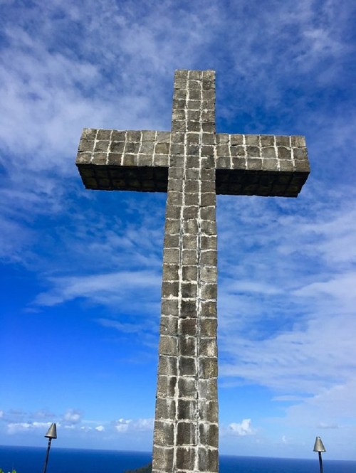 A large stone cross stands against a bright blue sky with scattered clouds, overlooking a distant ocean.