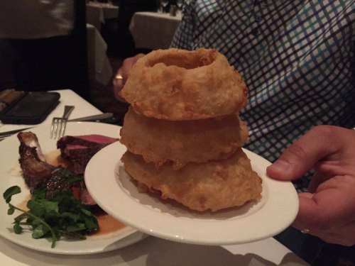 A person holds a plate with three stacked, golden-brown fried items, next to a plate of meat and greens.